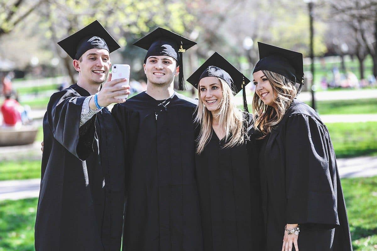 Four graduates in cap and gown taking selfie picture in park