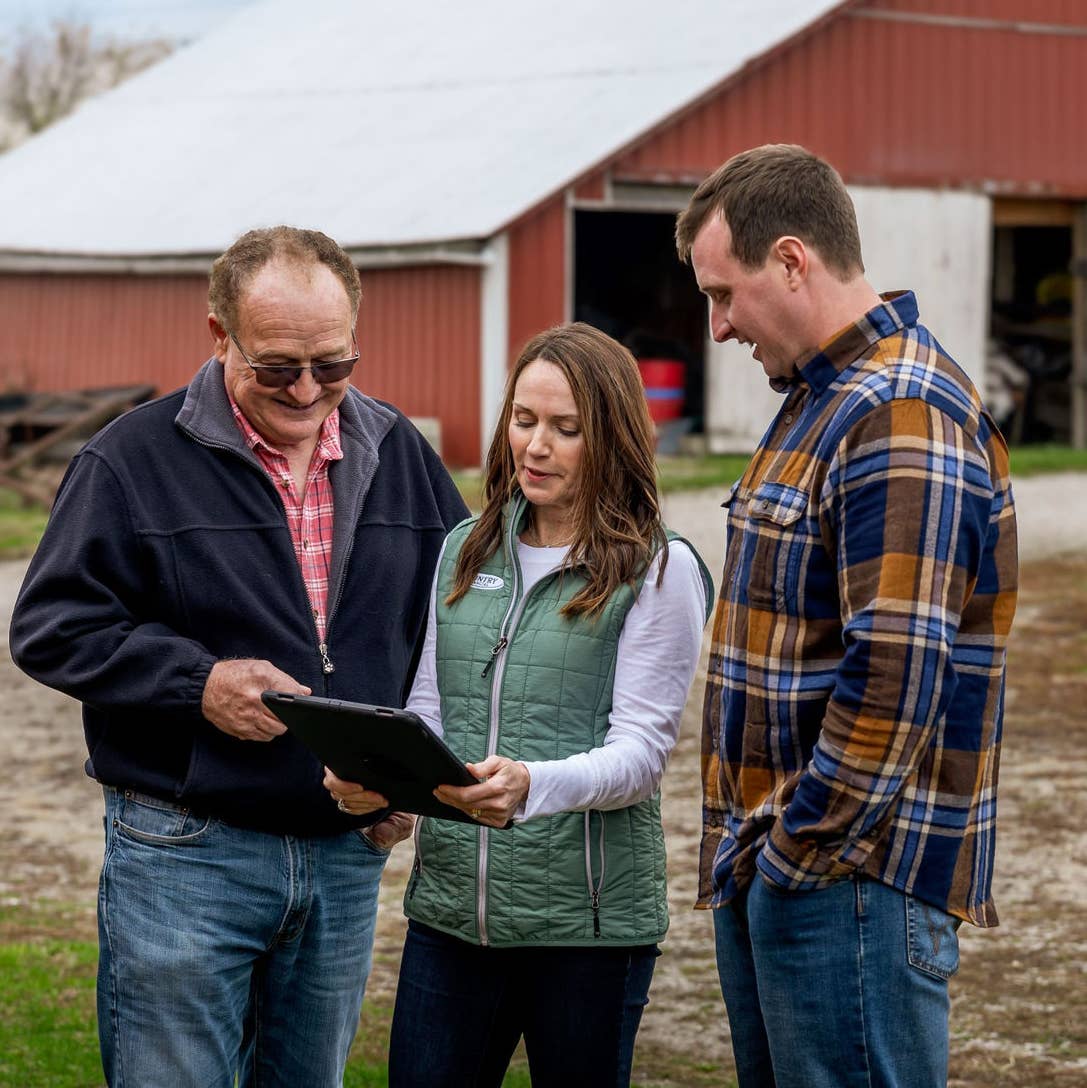A farm certified insurance rep looking at a tablet with two farm clients on their property