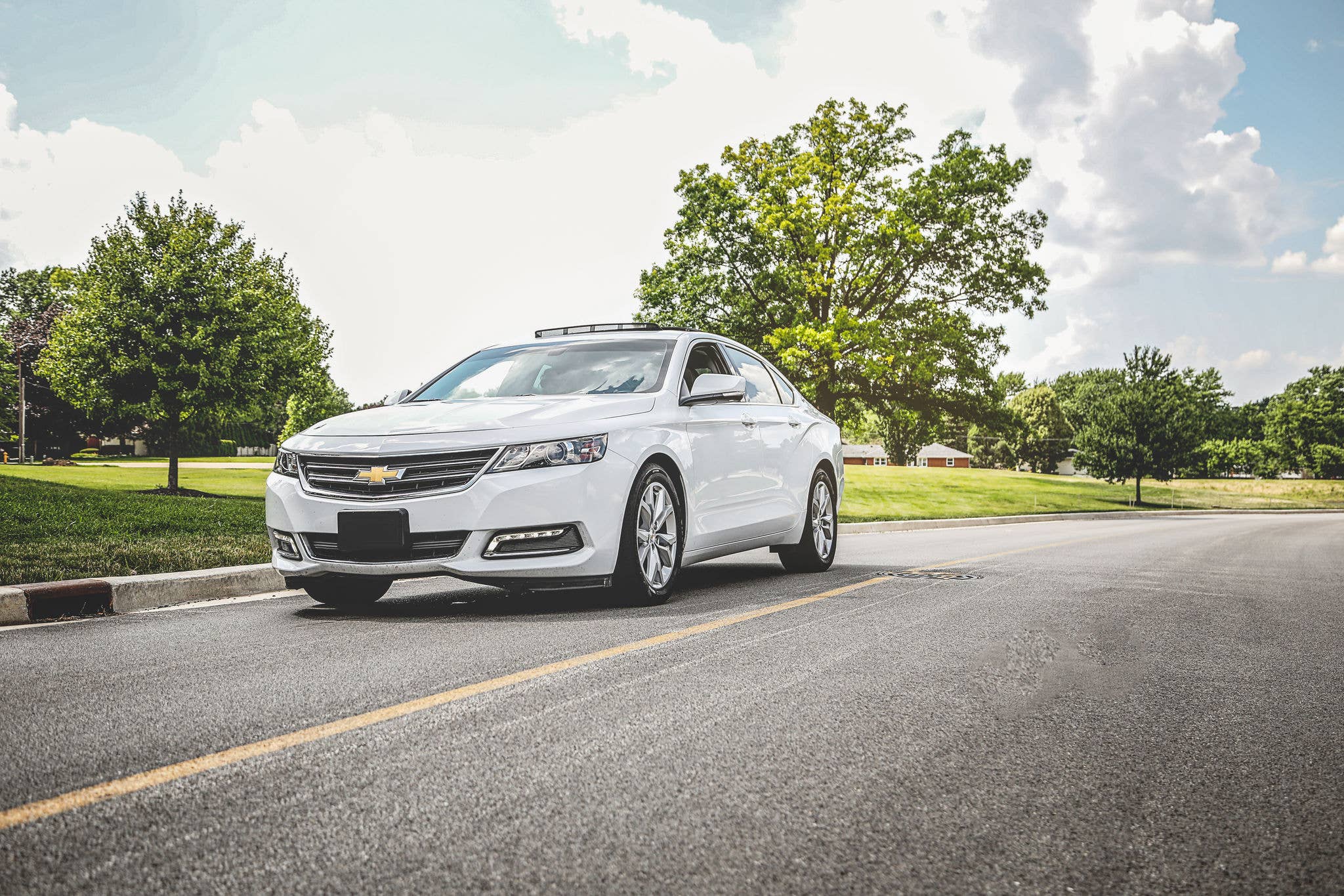 White Chevrolet driving on a road surrounded by trees and clear sky.